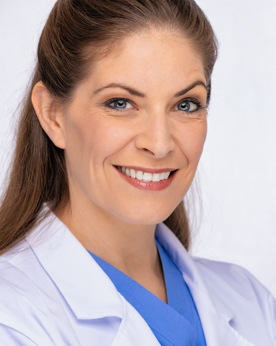 Cat as a warm medical professional in a white lab coat over blue scrubs, big genuine smile